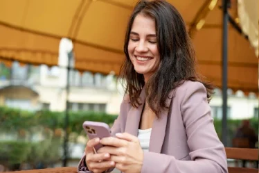 Mulher sorri ao mexer no celular sentada na rua