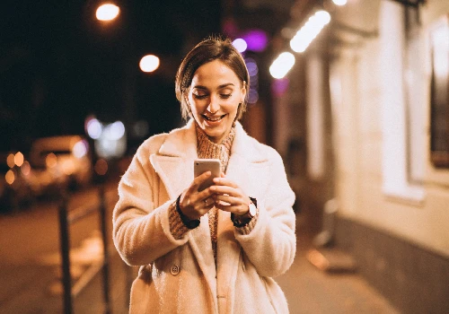 Mulher sorri mexendo no celular enquanto caminha na rua